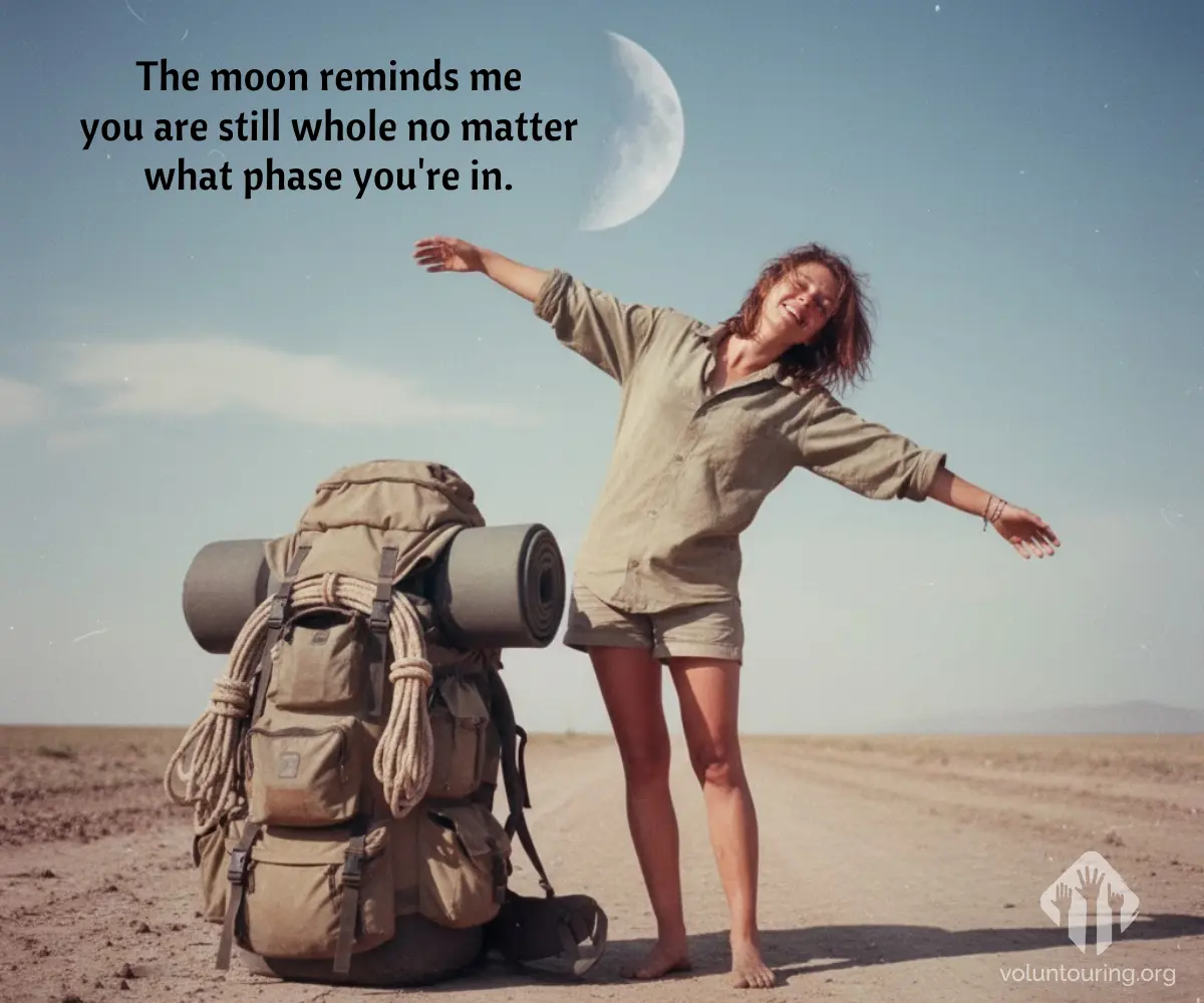 Happy barefoot traveler with arms outstretched beside a large backpack on a desert road, crescent moon visible in a clear blue sky. Quote reads, “The moon reminds me you are still whole no matter what phase you’re in.”