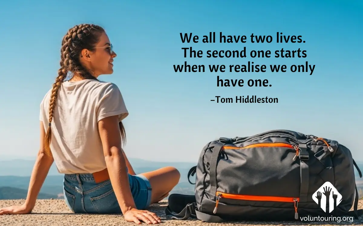A young woman with braided hair sits on a mountain viewpoint next to a travel backpack, gazing into the distance under a clear blue sky. A motivational quote is overlaid: 'We all have two lives. The second one starts when we realise we only have one.' – Tom Hiddleston. Voluntouring.org logo is visible in the corner.