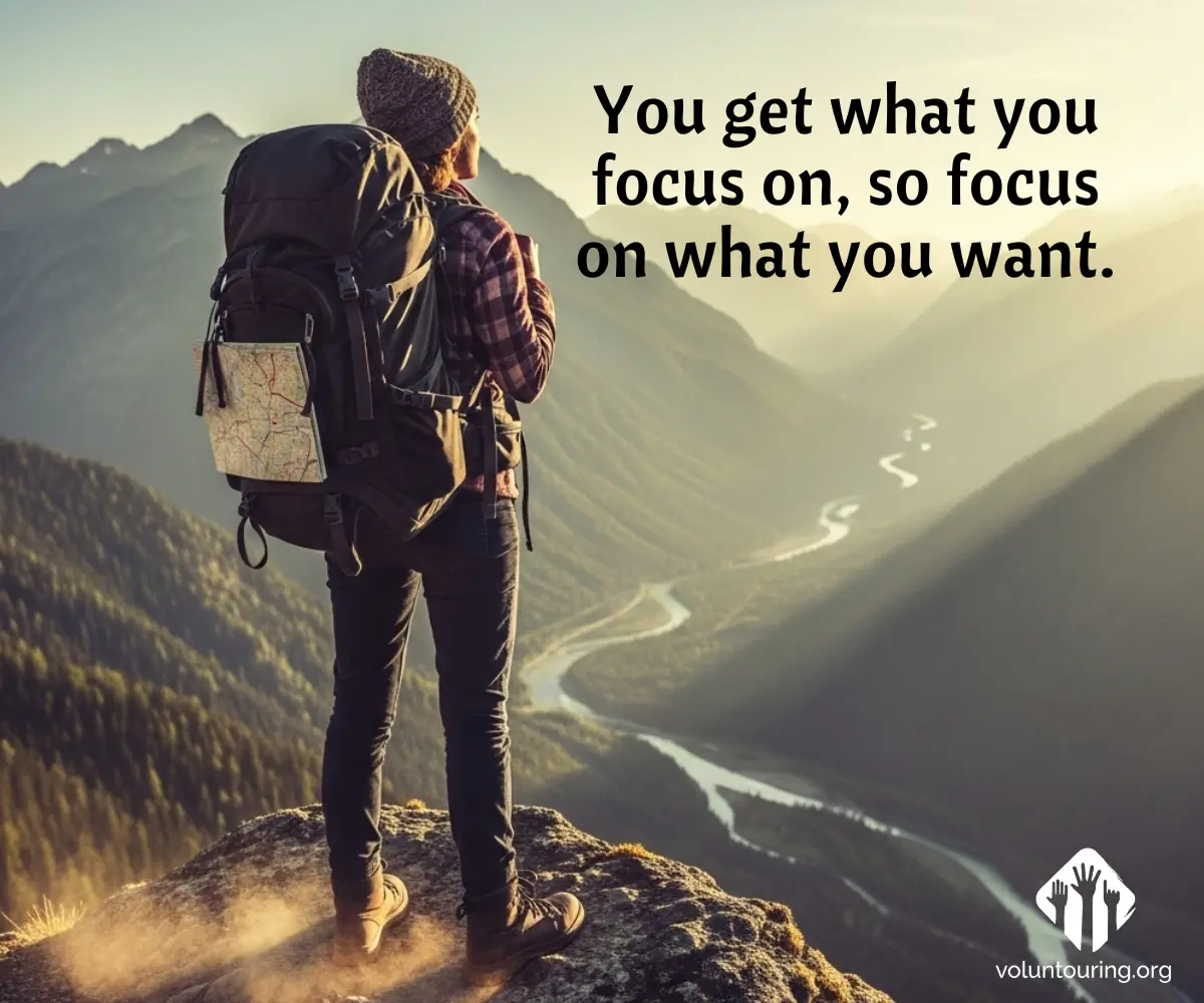 Hiker with backpack standing on mountain peak overlooking winding river valley at sunrise with motivational quote 'You get what you focus on, so focus on what you want' – Voluntouring.org