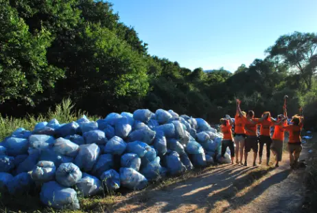 Psyclean volunteers in orange shirts celebrating next to a large pile of collected trash bags after a successful cleanup mission in a forested area.