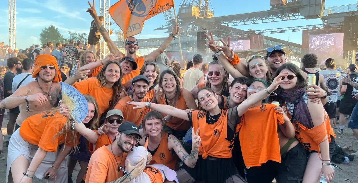 Group of Psyclean volunteers in orange shirts celebrating together at a European electronic music festival, smiling and posing in front of the stage with an orange crew flag.