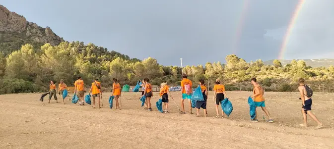Psyclean volunteers in orange shirts walking across a festival site with blue trash bags under a rainbow sky, cleaning up a natural landscape.