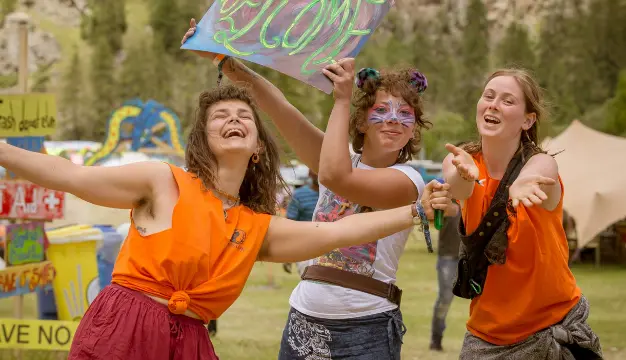 Three cheerful Psyclean volunteers in orange shirts welcome festival guests with open arms and painted signs at a colorful eco-conscious music event.