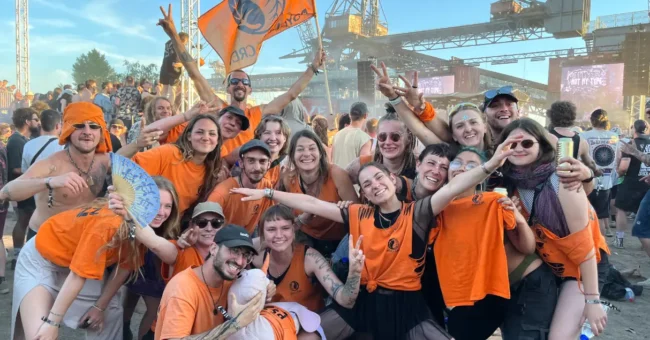 Group of Psyclean volunteers in orange shirts celebrating together at a European electronic music festival, smiling and posing in front of the stage with an orange crew flag.