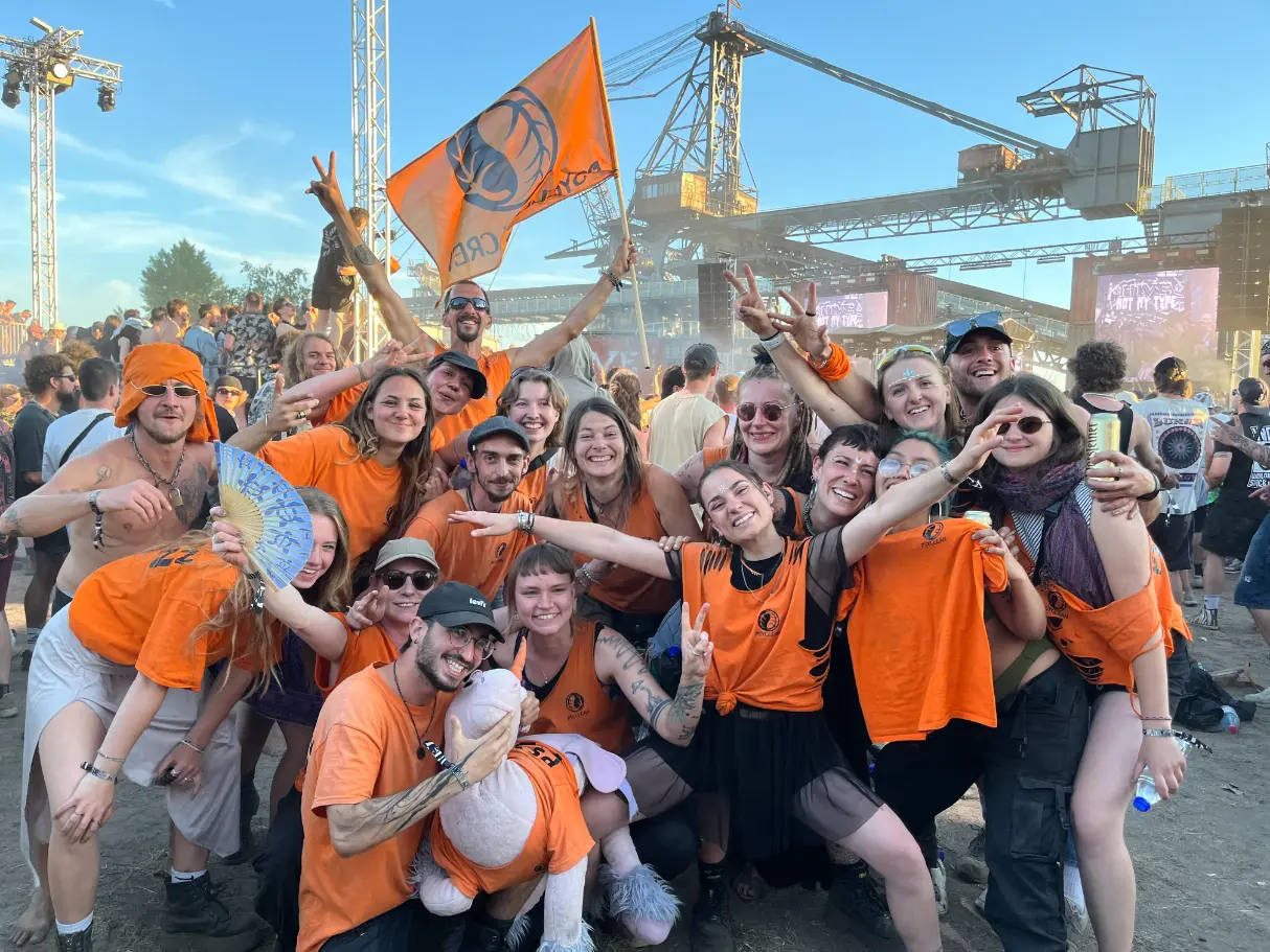 Group of Psyclean volunteers in orange shirts celebrating together at a European electronic music festival, smiling and posing in front of the stage with an orange crew flag.
