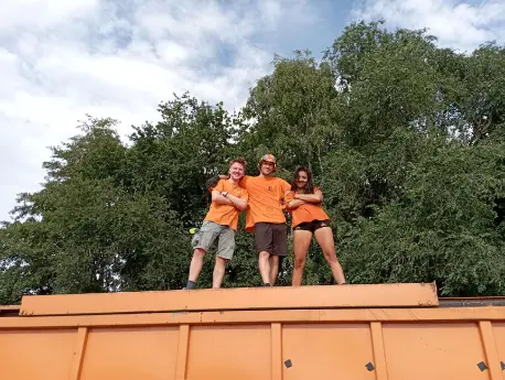 Three Psyclean volunteers in orange shirts posing confidently on top of a large orange container, with green trees and a blue sky in the background.
