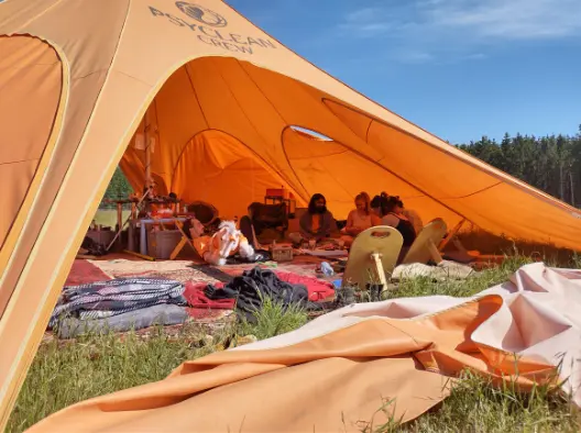 Volunteers relaxing inside the Psyclean Crew community tent at a European festival, surrounded by cushions, blankets, and shade under an orange canopy.