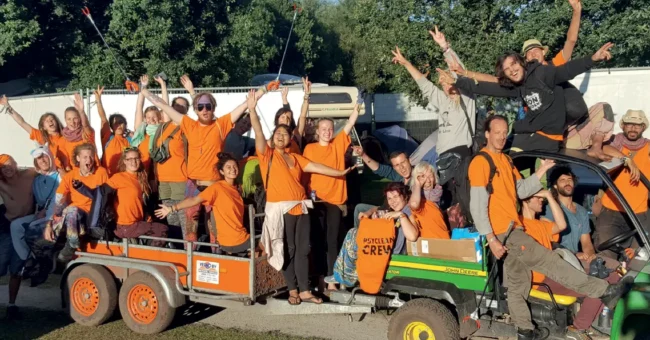 Group photo of Psyclean crew volunteers in bright orange shirts celebrating at a European music festival cleanup mission.