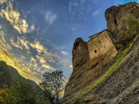 A dramatic view of the Castello della Pietra (Castle of the Stone) built into a rocky cliffside in Vobbia, Liguria, Italy, at sunset.