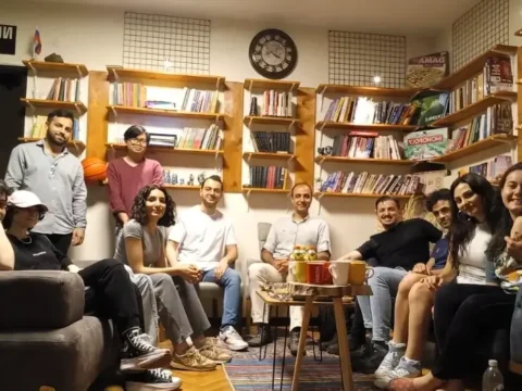 Group of international volunteers and local students socializing in a cozy English learning center in Istanbul, surrounded by bookshelves and cultural decor.