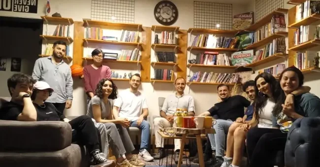 Group of international volunteers and local students socializing in a cozy English learning center in Istanbul, surrounded by bookshelves and cultural decor.