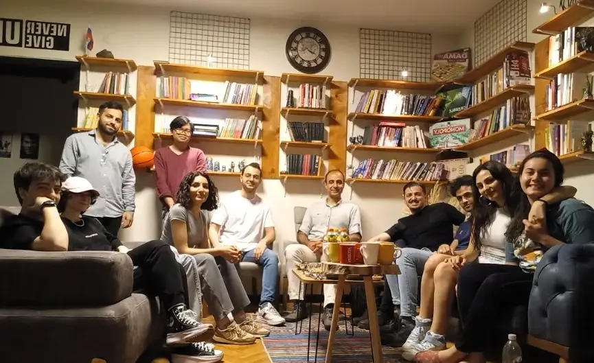 Group of international volunteers and local students socializing in a cozy English learning center in Istanbul, surrounded by bookshelves and cultural decor.