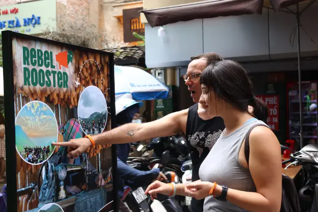 Two volunteers looking at a promotional board of Rebelrooster Hostel in Hanoi, Vietnam, featuring images of local culture, landscapes, and community activities