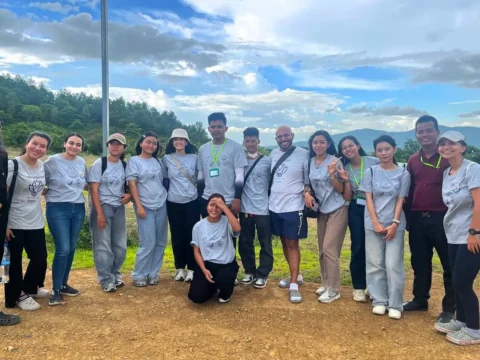 Group of international and Cambodian volunteers smiling outdoors in Kampot, Cambodia, during a community activity with AHHA Education, wearing matching grey t-shirts and ID badges with hills and trees in the background.