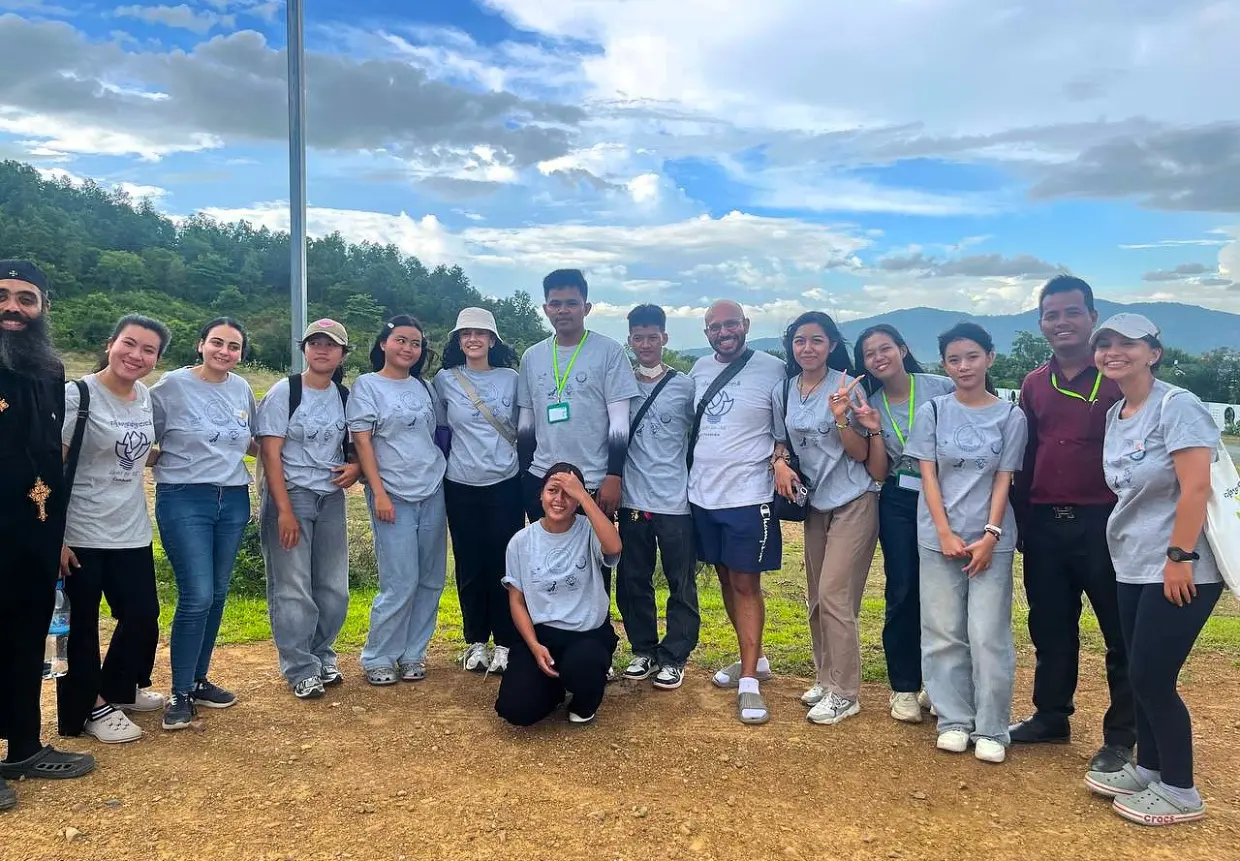 Group of international and Cambodian volunteers smiling outdoors in Kampot, Cambodia, during a community activity with AHHA Education, wearing matching grey t-shirts and ID badges with hills and trees in the background.