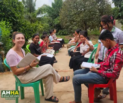 International volunteer teaching Cambodian students outdoors at AHHA Education in Kampot, surrounded by greenery. Students are holding books and smiling while engaging in an English learning activity.