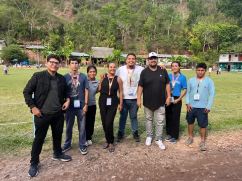 Group of Fundación Territorial Shamuy volunteers and staff standing on a grassy field in Tarapoto, Peruvian Amazon, with rainforest village buildings and people in the background.
