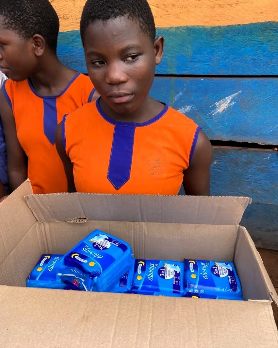 Girl in an orange school uniform in Jinja, Uganda standing beside a box of donated sanitary pads provided by Mothers and Children Uganda to support girls’ menstrual health and education.