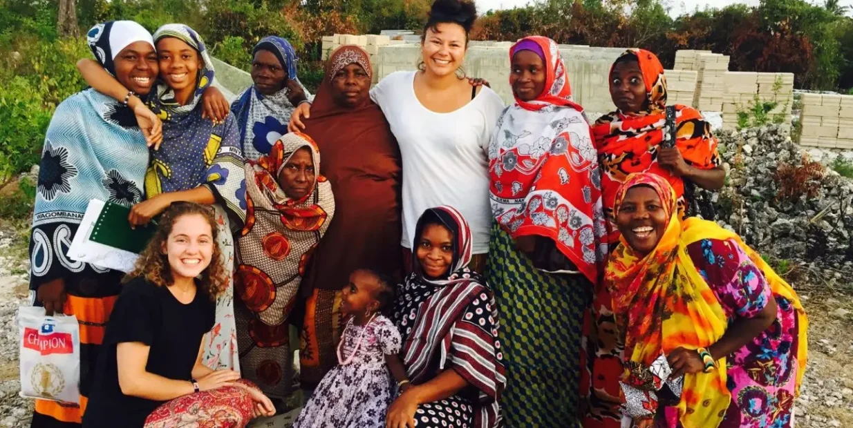 A smiling group of PLANE Uganda volunteers with local women and a child in Fort Portal, wearing colorful patterned clothing and standing outdoors during a community project.