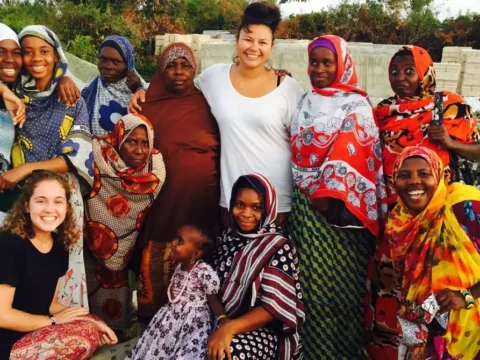 A smiling group of PLANE Uganda volunteers with local women and a child in Fort Portal, wearing colorful patterned clothing and standing outdoors during a community project.