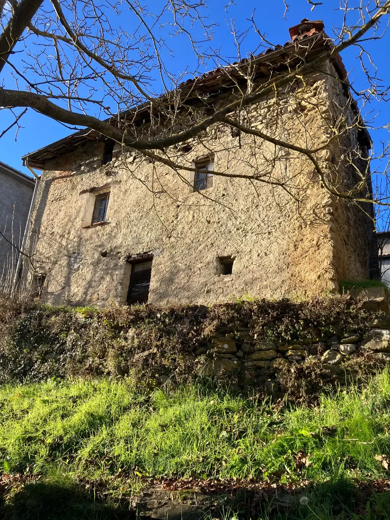 Rustic stone house in Cairo Montenotte, Liguria, Italy, being restored for the ColivingLiguria volunteer and remote work project, with a sunny façade, grassy foreground, and bare tree branches against a clear blue
