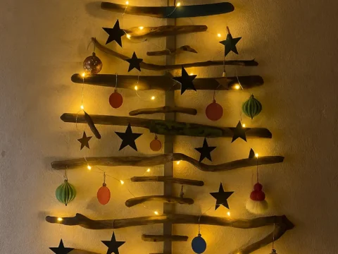 Wall mounted Christmas tree made from recycled driftwood and reclaimed wood, decorated with handmade paper ornaments and warm yellow fairy lights on a white plaster wall.