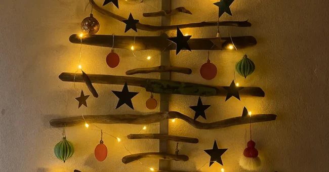 Wall mounted Christmas tree made from recycled driftwood and reclaimed wood, decorated with handmade paper ornaments and warm yellow fairy lights on a white plaster wall.