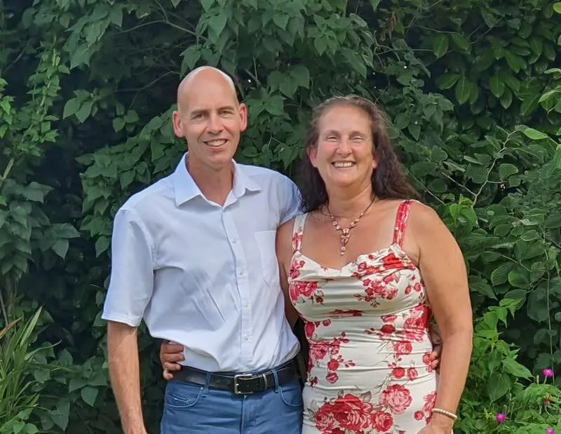 Host couple for a volunteer exchange in Vinaros, Castellon, Spain, standing outdoors in front of green foliage and smiling at the camera.