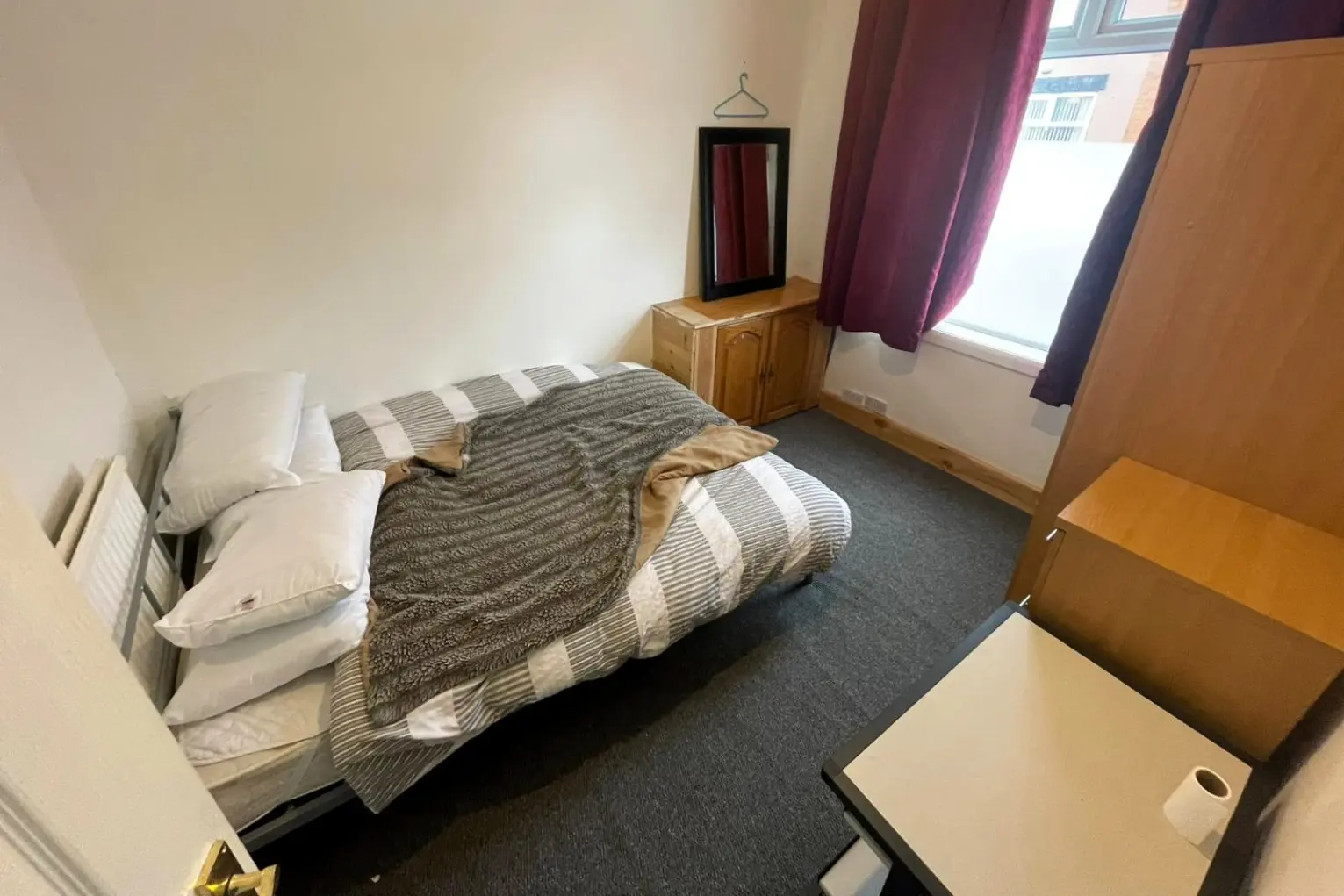 Simple private bedroom in a Manchester home with a double bed, wardrobe, small desk and window with dark curtains, offered as volunteer accommodation.