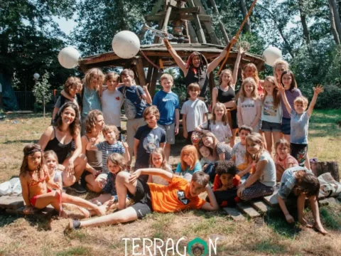 Group of children and camp staff posing outdoors in a wooded area in front of a wooden play structure with white balloons. “Terragon camps” text logo appears at the bottom of the image.