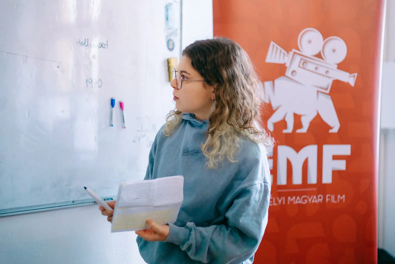 Volunteer holding notes and a pen while looking at a whiteboard during a session, with a Transylvanian Hungarian Films Association (THF) banner in the background.