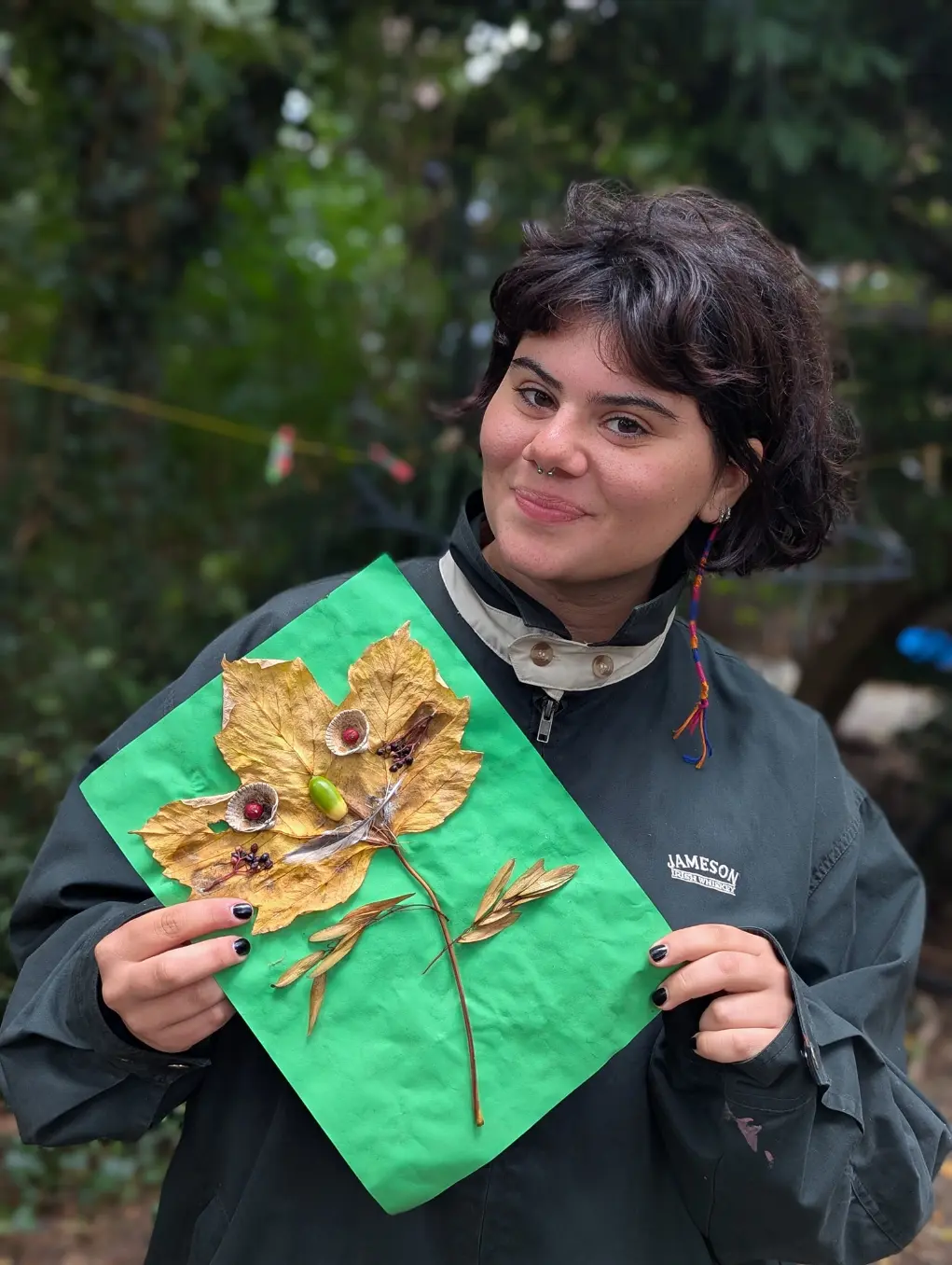 Close-up of an adult participant at a nature camp holding a green sheet with a leaf art collage made from dried leaves, acorns, seeds, and small natural items, photographed outdoors in a wooded setting in Amsterdam, Netherlands.