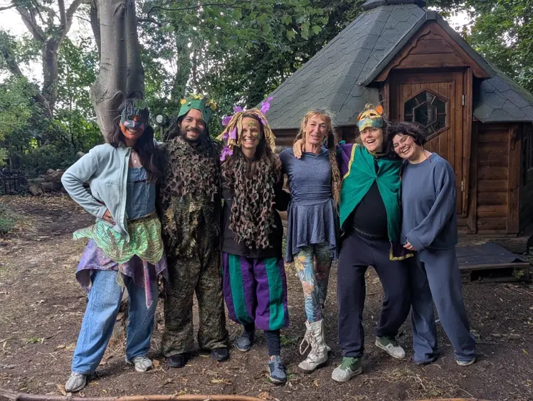 Group of adult participants standing together outdoors in front of a small wooden cabin in a forest setting, wearing colorful and playful costumes during a nature camp activity in Amsterdam.
