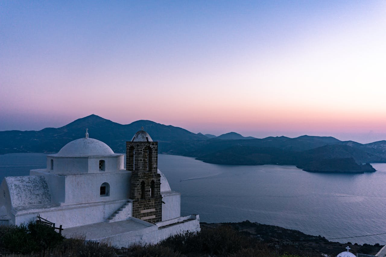 Whitewashed Greek Orthodox church with a stone bell tower overlooking the Aegean Sea at sunset on Milos, Greece.