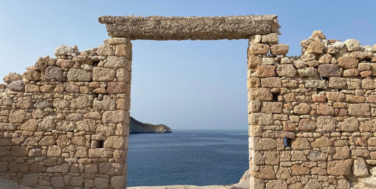 Stone doorway in a rugged old wall framing a view of the blue sea and cliffs on Milos island, Cyclades, Greece.