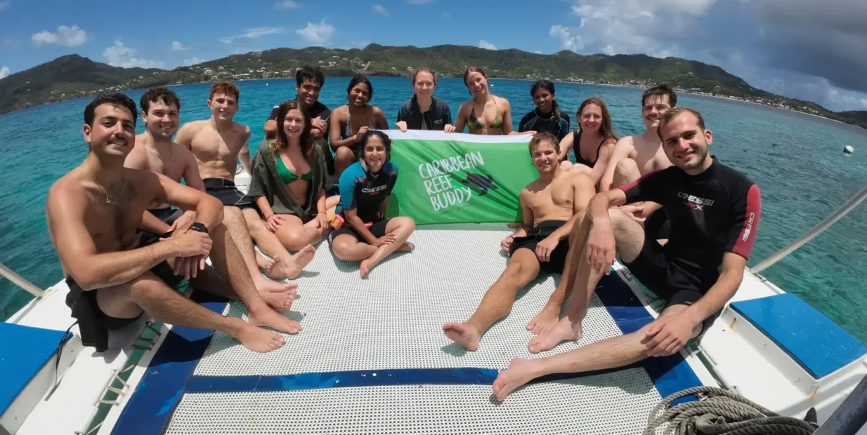 Group of marine conservation volunteers sitting on a boat and holding a Reef Buddy banner above clear turquoise sea, with a tropical coastline in the background.