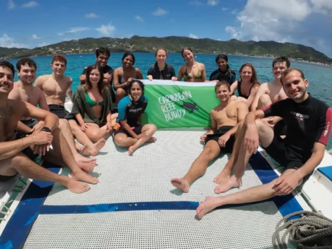 Group of marine conservation volunteers sitting on a boat and holding a Reef Buddy banner above clear turquoise sea, with a tropical coastline in the background.