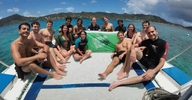 Group of marine conservation volunteers sitting on a boat and holding a Reef Buddy banner above clear turquoise sea, with a tropical coastline in the background.