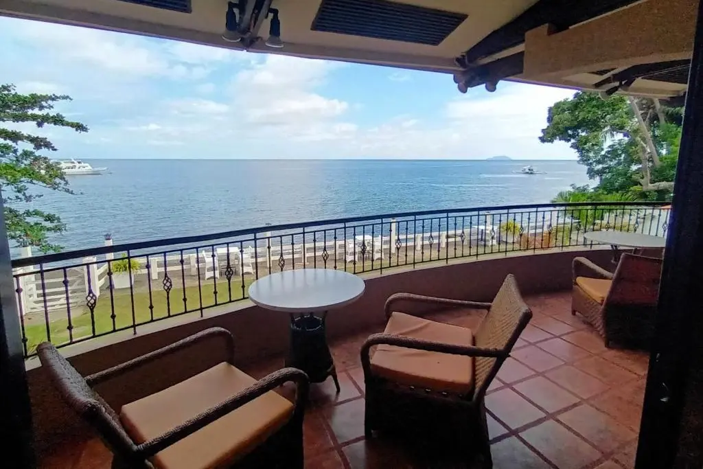 Covered balcony terrace with wicker chairs and a small table overlooking a calm ocean, beach, and boats, with a distant island on the horizon.