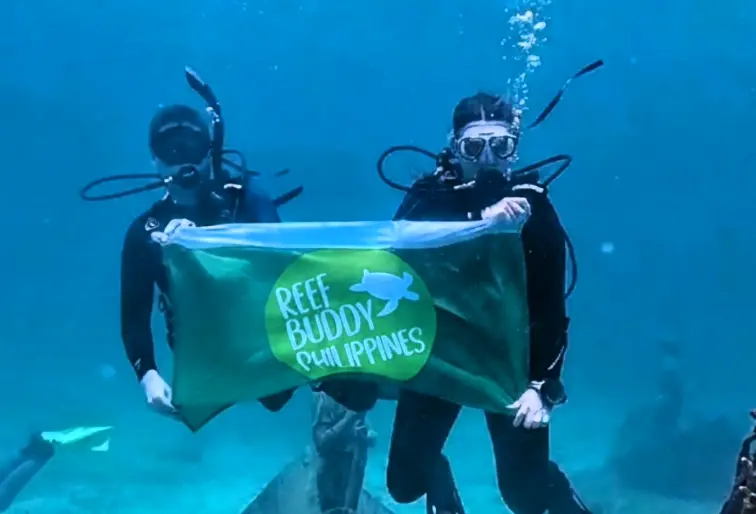 Two scuba divers underwater holding a Reef Buddy Philippines banner, with bubbles rising in clear blue water.