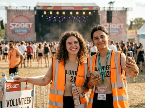 Two Sziget Festival volunteers in orange high-visibility vests smiling near the main stage, with festival crowd, staff passes, and volunteer area in the background