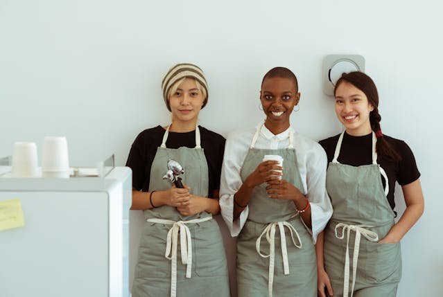 Three young people wearing aprons standing together in a shared work space, representing community life and teamwork during a volunteer exchange project.