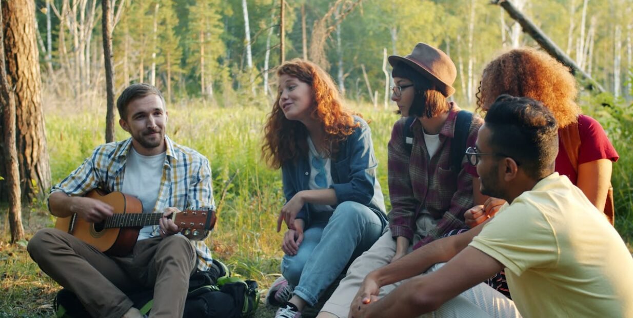A group of young people sitting in a forest during a summer camp, sharing music and conversation with a guitar, representing community life, creativity and outdoor volunteering in Saverne, France.