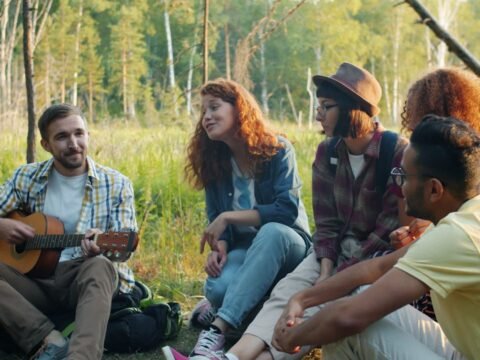 A group of young people sitting in a forest during a summer camp, sharing music and conversation with a guitar, representing community life, creativity and outdoor volunteering in Saverne, France.