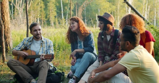 A group of young people sitting in a forest during a summer camp, sharing music and conversation with a guitar, representing community life, creativity and outdoor volunteering in Saverne, France.