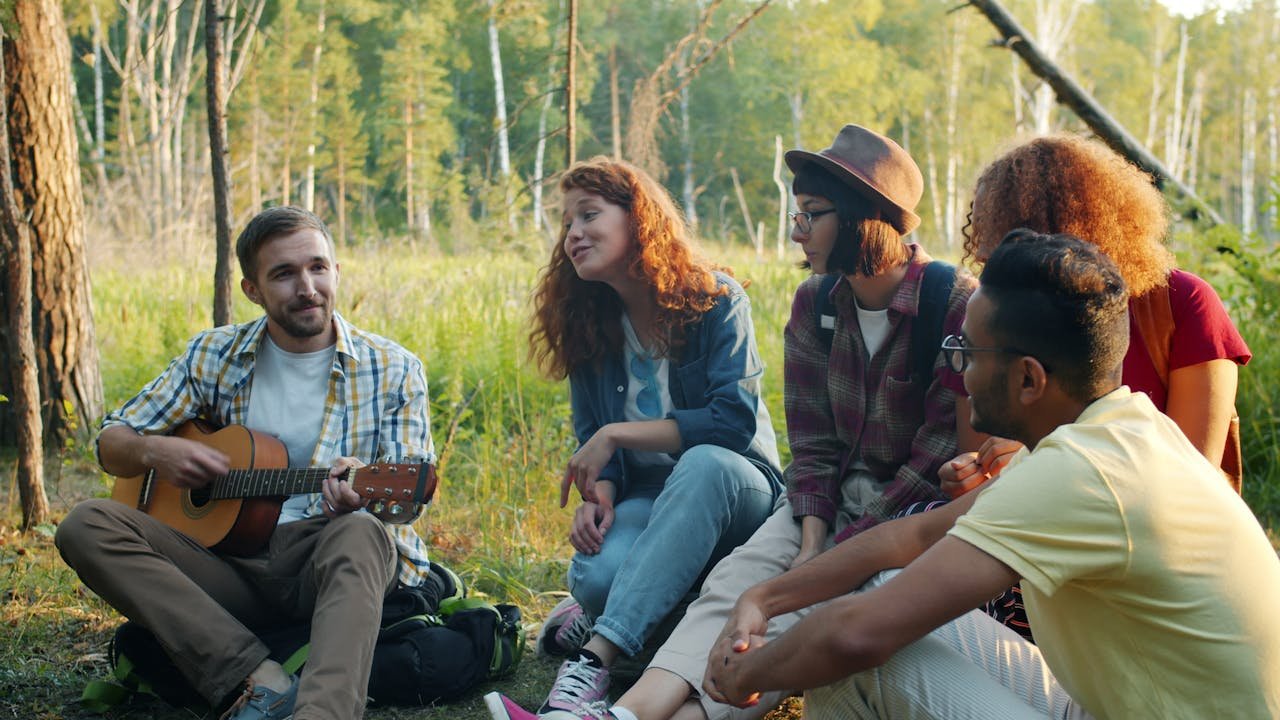 A group of young people sitting in a forest during a summer camp, sharing music and conversation with a guitar, representing community life, creativity and outdoor volunteering in Saverne, France.
