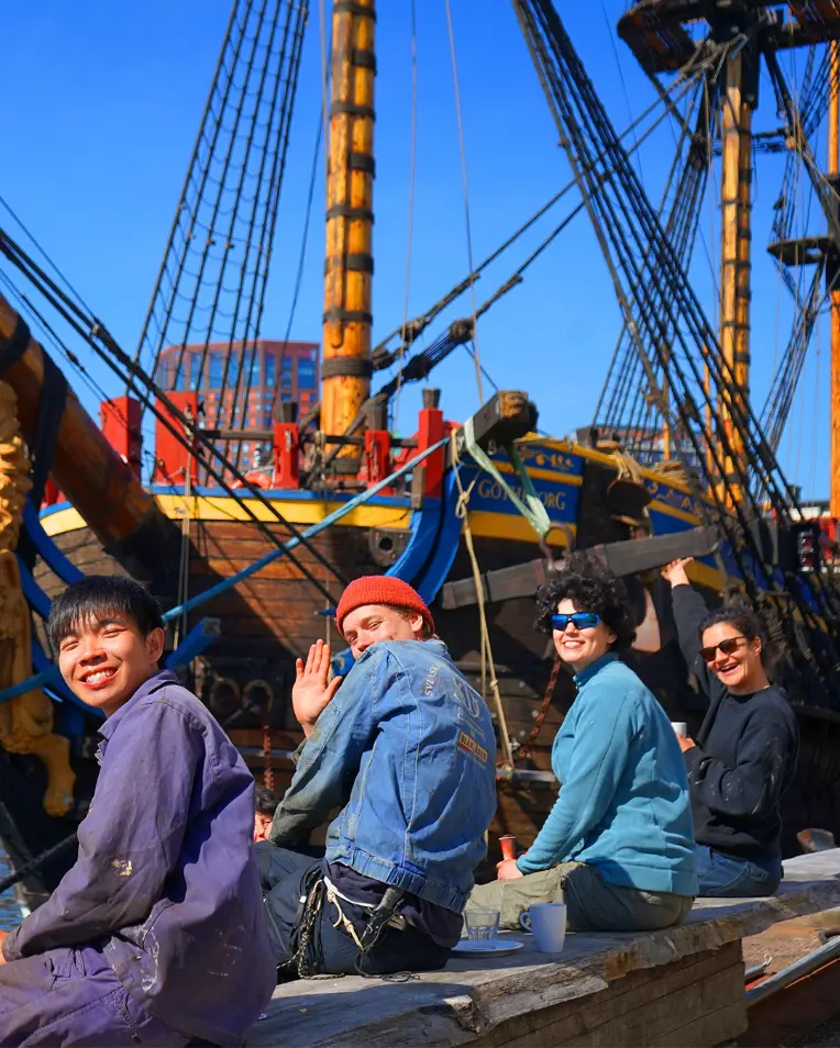 Smiling volunteers sitting beside the Götheborg wooden sailing ship in Gothenburg, Sweden, during a historic ship volunteering opportunity.