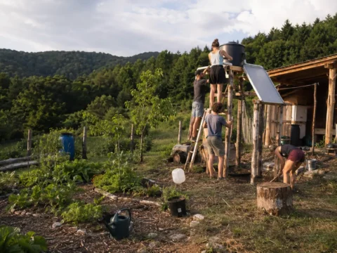 Young volunteers building a solar-heated garden shower beside a rustic wooden shelter in rural Hungary, surrounded by gardens, forested hills and an off-grid sustainability landscape.