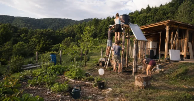 Young volunteers building a solar-heated garden shower beside a rustic wooden shelter in rural Hungary, surrounded by gardens, forested hills and an off-grid sustainability landscape.
