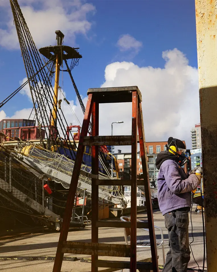 Volunteer wearing safety gear during hands-on maintenance work near the Götheborg historic wooden ship in Gothenburg, Sweden, for a ship volunteering opportunity.